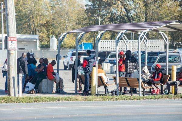 A group of migrant men gather around a bus stop.