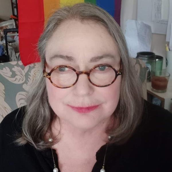 Woman with glasses sitting on a chair with a pride flag in the background.