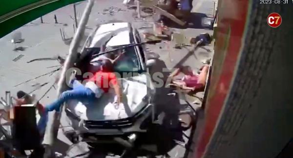 A man in a red t-shirt and blue jeans is pictured on top of a white SUV as debris falls beside him.