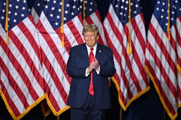 Do<em></em>nald Trump in a suit and tie standing in front of flags.