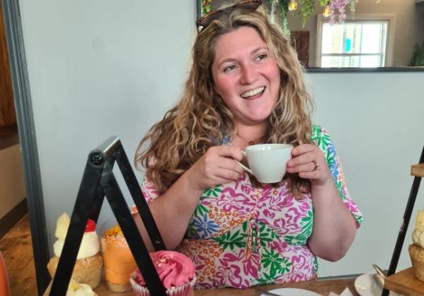 Emmie Harrison-West drinking a cup of tea during an afternoon tea out, with cakes on an afternoon tea stand in the foreground.