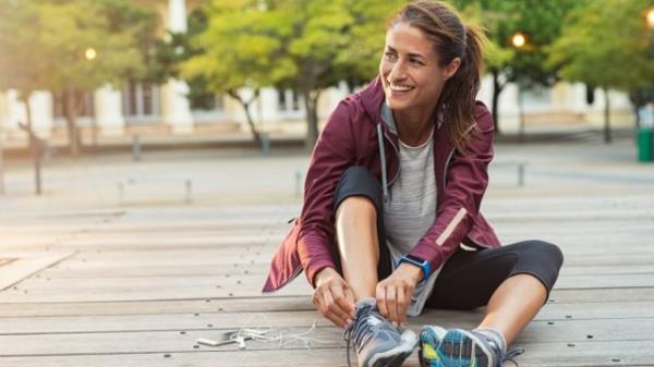 Mature fitness woman tie shoelaces on road. Cheerful runner sitting on floor on city streets with mobile and earpho<em></em>nes wearing sport shoes. Active latin woman tying shoe lace before running.