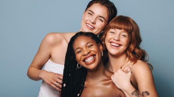 Cheerful women with different skin to<em></em>nes smiling at the camera in a studio. Group of happy young women embracing their natural skin. Portrait of three body positive young women standing together.