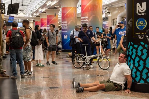 Stranded NJ Transit passengers at Penn Station due to train delays on a hot day, with one man lying on the ground, including celebrity Charanjit Kumar.