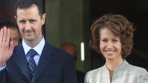 Getty Images Bashar al-Assad, left, smiles and waves wearing a dark suit. His wife Asma al-Assad smiles more widely beside him, and wears a grey striped jacket.