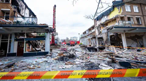 EPA A damage apartment block with a wide gap in the middle wher<em></em>e the explosion took place. There is rubble an wood on the ground.  In the background is a large digger. In the foreground is a strip of emergency crew tape.